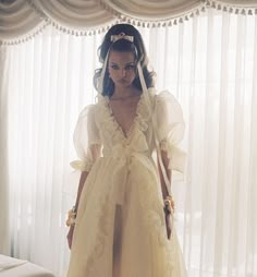 a woman in a yellow dress standing next to a window with sheer drapes on it