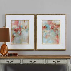 two framed paintings on the wall next to a table with a lamp and bookcase