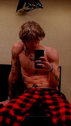 a shirtless man sitting on a bed looking at his cell phone while wearing red and black flannel