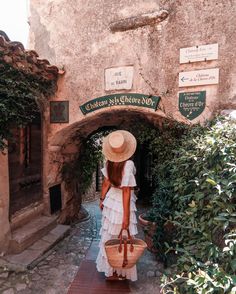 a woman in a white dress and straw hat carrying a wicker basket through an archway