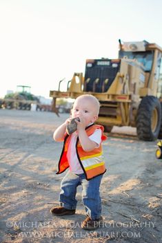 a little boy standing in the dirt with a construction vest on and holding something to his mouth