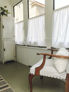 a white chair sitting next to a window in a room with two windows and a rug on the floor