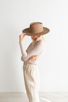 a woman in white pants and a brown hat is standing with her hands on her head