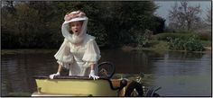 a woman in a white dress and hat on a yellow boat floating down a river