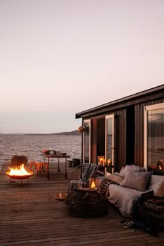 an outdoor fire pit on a wooden deck next to the ocean at dusk with chairs and tables around it