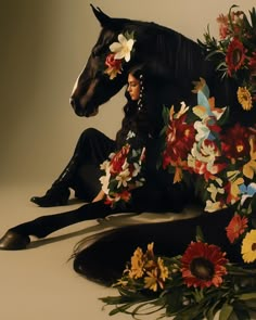 a woman sitting on the ground next to a black horse surrounded by flowers and plants