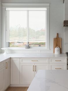 a kitchen with white cabinets and marble counter tops in front of a large open window