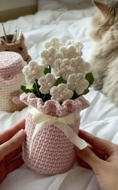 a person holding a small crocheted vase with flowers in it and a cat laying next to it