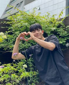 a young man making a heart shape with his hands while standing in front of some plants