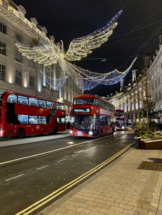 two red double decker buses driving down a street next to tall buildings with christmas lights on them