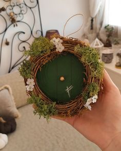 a hand holding a wreath with a green door and flowers on the inside, in front of a bed