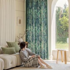 a woman sitting on the floor in front of a window with green drapes and curtains