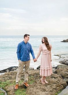 a man and woman holding hands while standing on rocks near the ocean