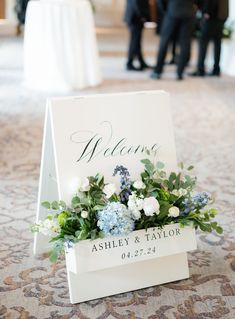 a welcome sign with flowers and greenery on the floor in front of people at a wedding