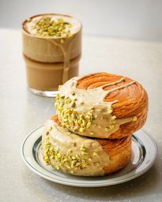 two croissants with frosting and pistachio sprinkles sit on a plate next to a cup of coffee