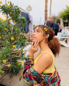 a woman standing next to a tree with yellow flowers on it's head and looking up at the sky