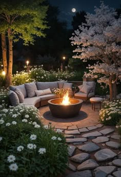 an outdoor fire pit surrounded by flowers and trees at night with lights on the ground