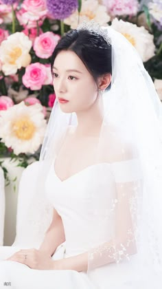 a woman in a white wedding dress sitting on a couch with flowers behind her and wearing a veil