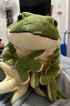 a stuffed frog sitting on top of a bed next to another stuffed animal in the background