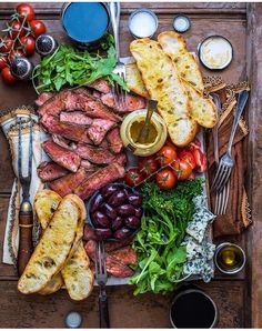 an assortment of meats, bread and vegetables on a wooden table with utensils