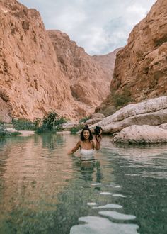 a woman standing in the middle of a river surrounded by rocky mountains and greenery