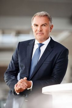 a man wearing a suit and tie standing in front of a white table with his hands folded