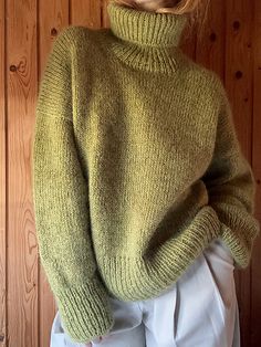 a woman standing in front of a wooden wall wearing a green sweater and white pants