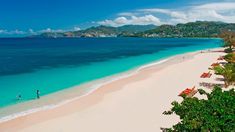 people are walking on the beach next to the blue water and clear waters, with umbrellas in the foreground