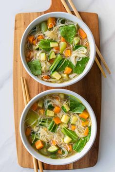 two bowls of vegetable noodle soup on a cutting board with chopsticks next to it