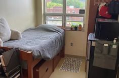 a small room with a bed, desk and refrigerator in it next to a window