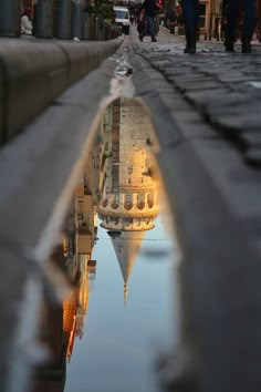 a reflection of a building in water on the ground with people walking around it and one person standing near by