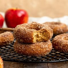 some sugary donuts are on a cooling rack with apples in the back ground