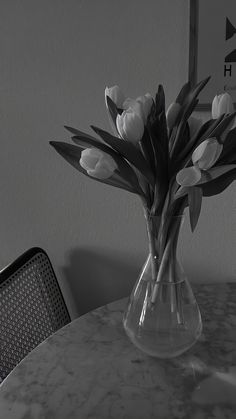 black and white photograph of flowers in vase on table with chair next to wall behind