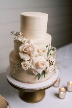 a wedding cake with white flowers and greenery sits on a table next to candles