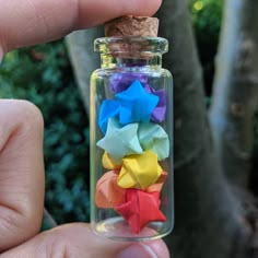 a hand holding a tiny glass bottle filled with colorful origami stars in front of a tree