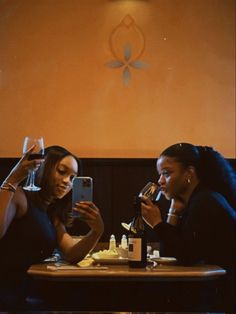 two women sitting at a table with wine glasses in front of them, looking at their cell phones