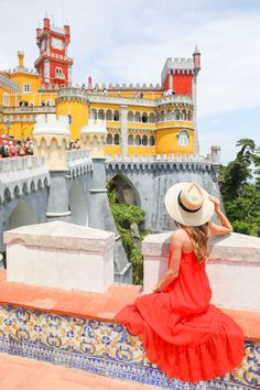 a woman in a red dress and hat sitting on a wall looking at the castle