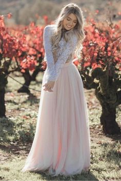 a woman wearing a long pink dress standing in front of some bushes and trees with red flowers