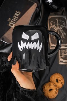 a hand holding a black coffee mug with white teeth on it and cookies next to it