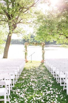 an outdoor ceremony set up with white chairs and flowers on the grass, near water