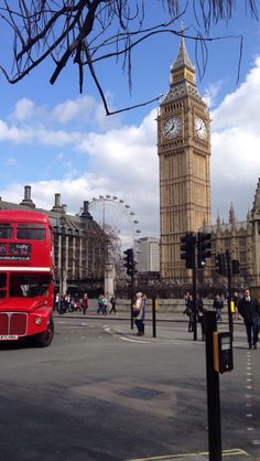 a red double decker bus driving down a street next to a tall tower with a clock on it's side