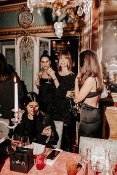 three women standing around a table with drinks and wine glasses in front of them at a party