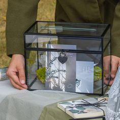 a person is holding a glass box with plants in it on top of a table