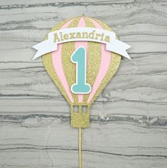 a pink and gold balloon cake topper with the number one on it's side