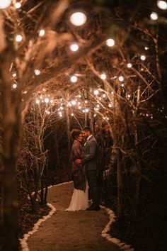 a bride and groom standing in the middle of a path surrounded by trees with lights