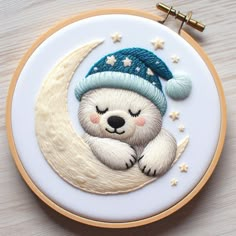 a white bear with a blue hat is sitting on the moon embroideryed onto a hoop