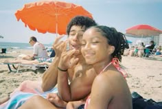 two people sitting on the beach under an orange umbrella and smiling at the camera man is holding his hand up to his face