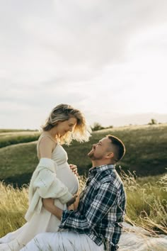 a pregnant woman sitting next to a man on top of a grass covered field in the sun