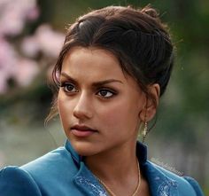 a close up of a person wearing a blue shirt and gold necklace with flowers in the background