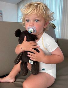 a little boy sitting on a couch holding a teddy bear with a pacifier in his mouth